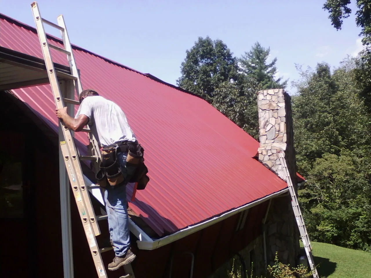 Roofer on a ladder beside a red metal roof and stone chimney — Soffit & Fascia Repair by Roofing Brothers in Barryville